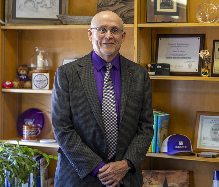 A man in a suit poses for a portrait in his office and in front of a wooden bookshelf.