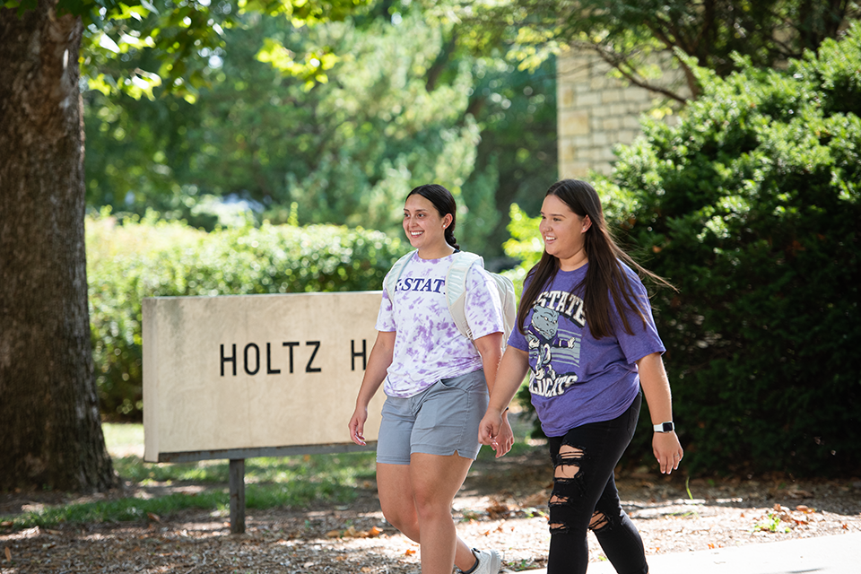 Two female college students walk on campus on a sunny day in front of a limestone sign that says "Holtz Hall.