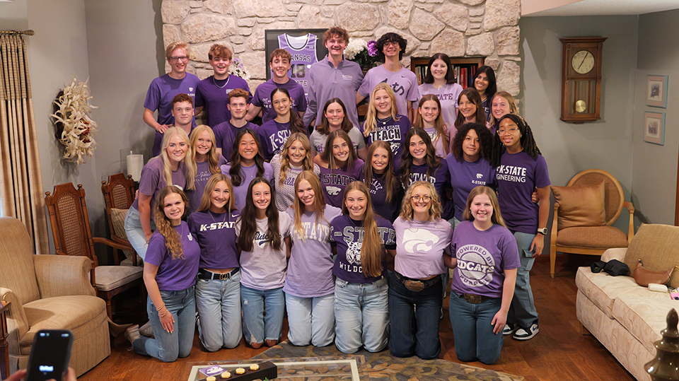 A group of college students in purple clothing poses for a group portrait in a large living room area with a chimney.