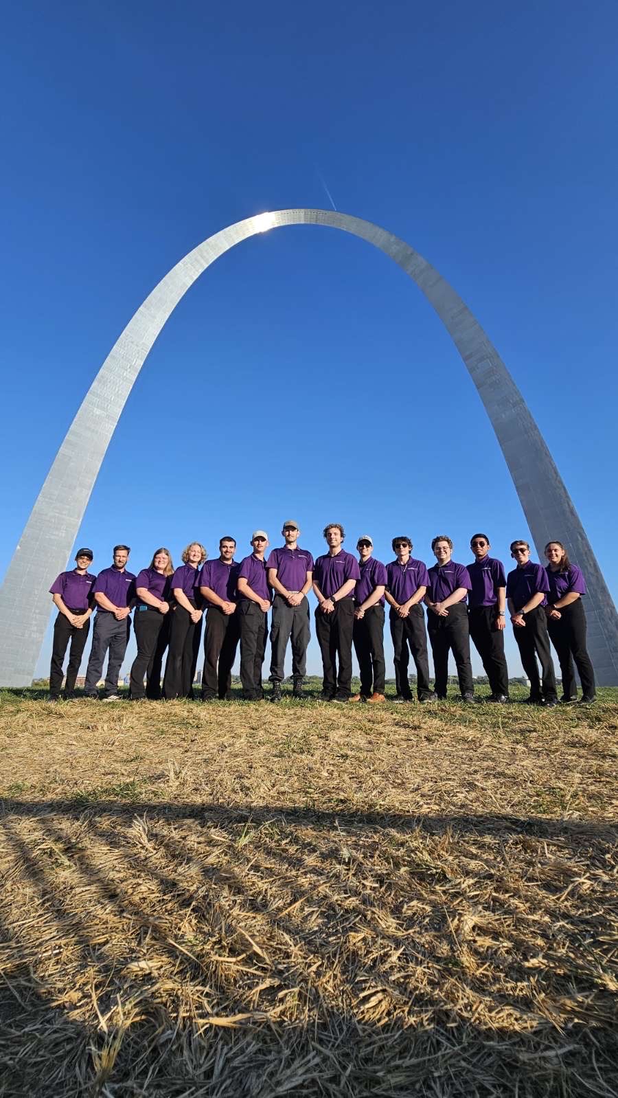 A group of 14 students in purple polos and black pants stand below the St. Louis Arch with a blue sky in the background and a field in the foreground.