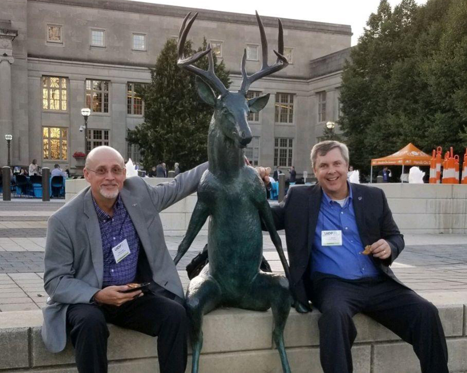 Paul Lowe and a fellow research professional sit on an outdoor bench with a sculpture of a buck.