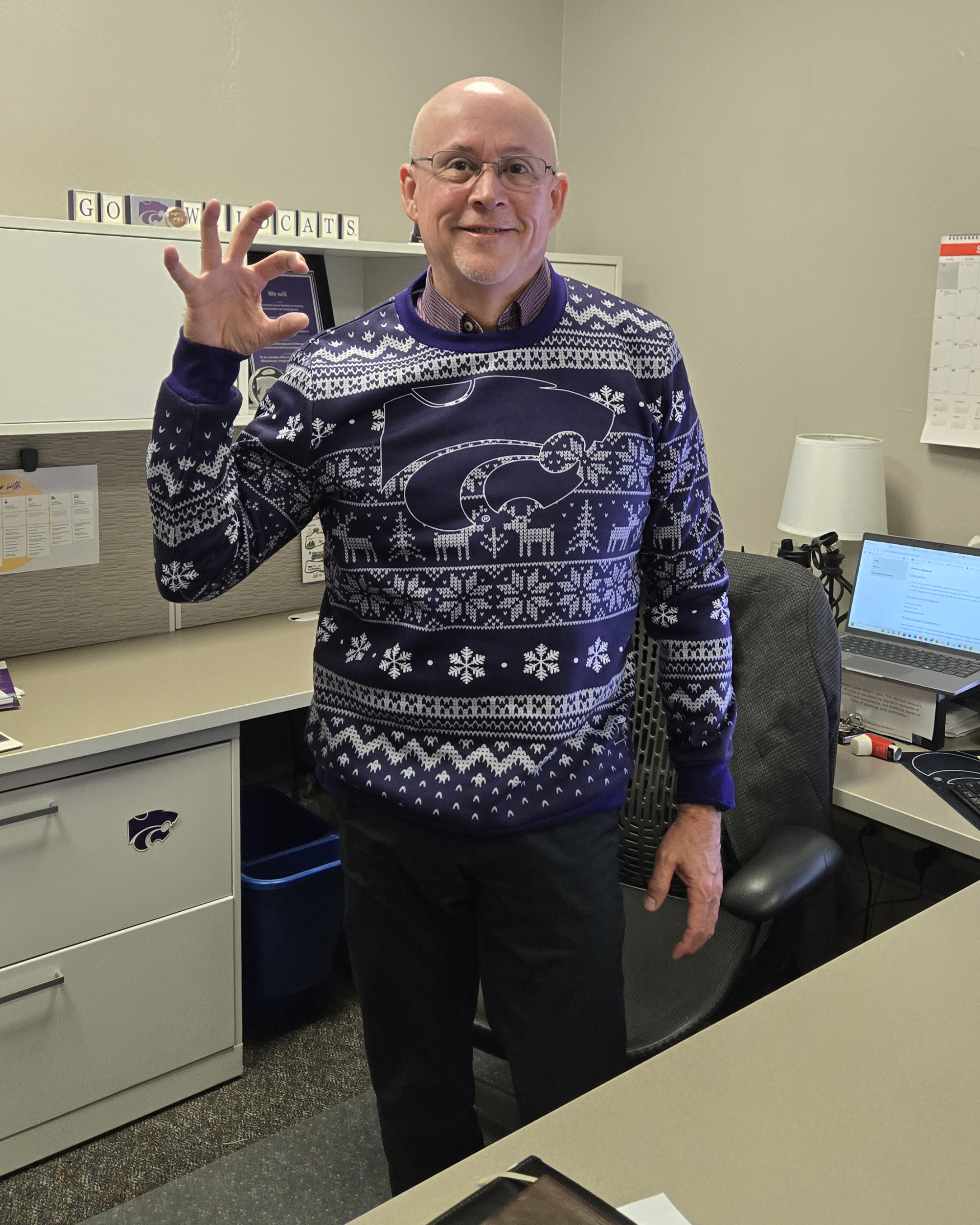 Paul Lowe wears a purple holiday sweater, flashes a W-C hand symbol and poses for a portrait in his office.