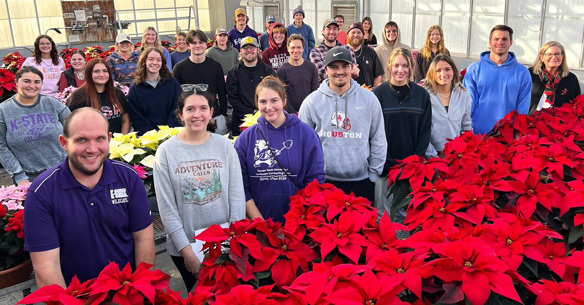 About 30 people, a mix of students and gardens volunteers, stand in a greenhouse filled with red and pink poinsettias.